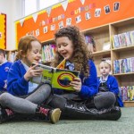 Children sitting on the floor of a library reading a colourful book together, with other children browsing shelves in the background.