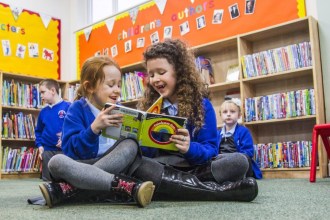 Children sitting on the floor of a library reading a colourful book together, with other children browsing shelves in the background.
