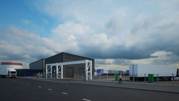 A sleek, single-story industrial building with dark grey cladding and large glass windows. Behind it rises a taller, curved structure with horizontal silver panelling. The scene is bright with a clear blue sky and scattered white clouds.