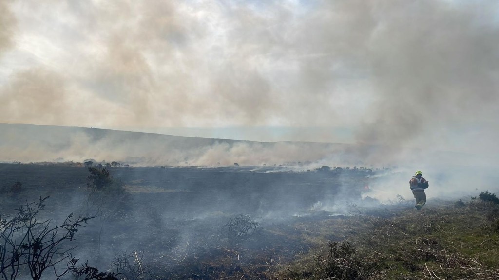 A firefighter in high-visibility gear walking across smoke-covered, fire-blackened ground at Cefn Bryn in Gower, with smouldering vegetation stretching across the hillside behind them.