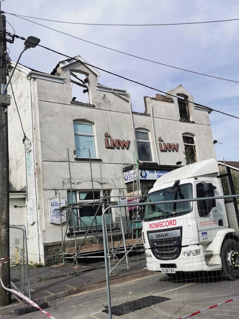 The fire-damaged former LNW Club building on Sterry Road in Gowerton, showing the gutted roofline and scaffolding erected across the front, with a Rowecord DAF lorry parked outside and barrier tape across the road.