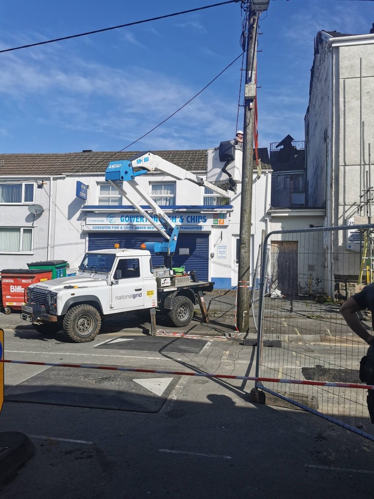 A National Grid cherry picker and Defender vehicle on Sterry Road in Gowerton as engineers work on overhead power lines, with the blue shopfront of Gowerton Fish and Chips visible to the left and the fire-damaged LNW building to the right.