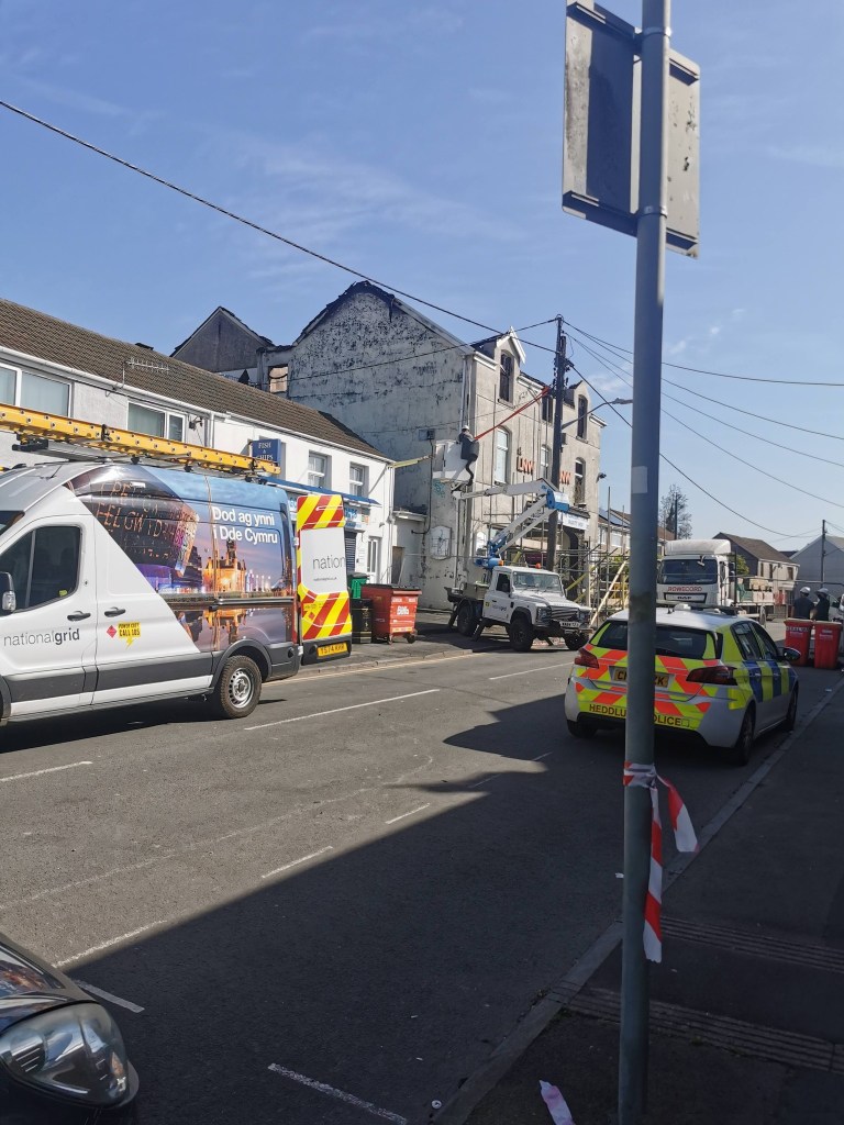 A wide view of Sterry Road in Gowerton showing a South Wales Police car, National Grid van, and a cherry picker working on overhead power lines, with the fire-damaged former LNW Club building visible in the background.