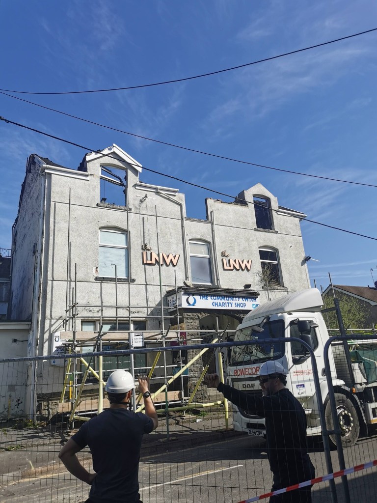 Two workers wearing hard hats standing outside a metal security barrier on Sterry Road in Gowerton, looking up at the gutted former LNW Club building with scaffolding across the front facade under a bright blue sky.