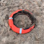 A badly burned orange lifebuoy ring lying partially buried in sand on Aberavon Beach, its foam interior destroyed by fire