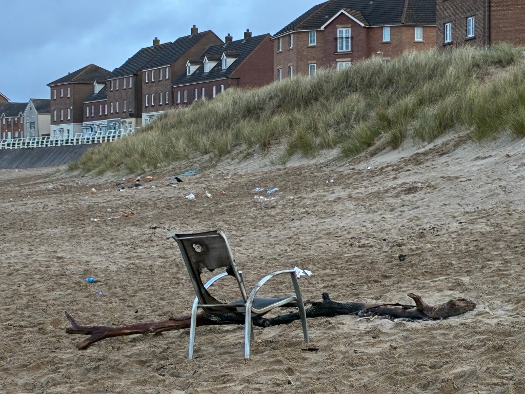 A burned metal garden chair and charred pieces of wood left on the sand at Aberavon Beach, with litter scattered across the beach in front of a row of residential houses and sand dunes