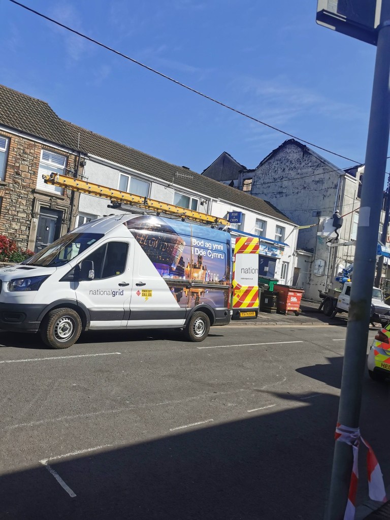 A National Grid van on Sterry Road in Gowerton with the burned-out gable end of the former LNW Club visible in the background under a blue sky, with barrier tape and emergency vehicles also visible.