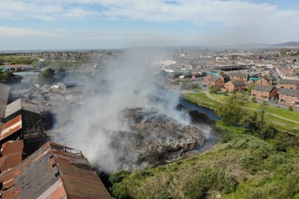 Aerial drone view showing a large fire still smouldering at an industrial site in Port Talbot, with a thick plume of grey smoke rising above the ruins of a collapsed building alongside a canal, with residential streets and the wider town visible in the background