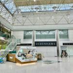 The interior of the Quadrant Shopping Centre in Swansea showing the vacant former Debenhams unit, with the Debenhams fascia still visible across three entrances, a Krispy Kreme kiosk in the foreground and shoppers passing through the atrium beneath a glass roof