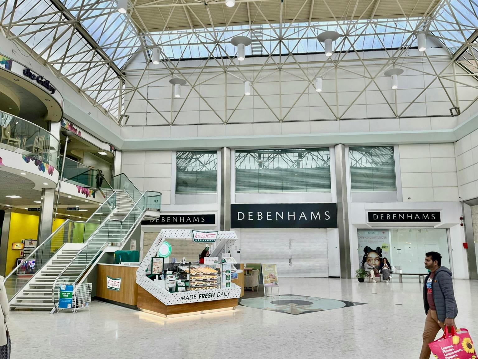 The interior of the Quadrant Shopping Centre in Swansea showing the vacant former Debenhams unit, with the Debenhams fascia still visible across three entrances, a Krispy Kreme kiosk in the foreground and shoppers passing through the atrium beneath a glass roof