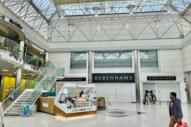 The interior of the Quadrant Shopping Centre in Swansea showing the vacant former Debenhams unit, with the Debenhams fascia still visible across three entrances, a Krispy Kreme kiosk in the foreground and shoppers passing through the atrium beneath a glass roof