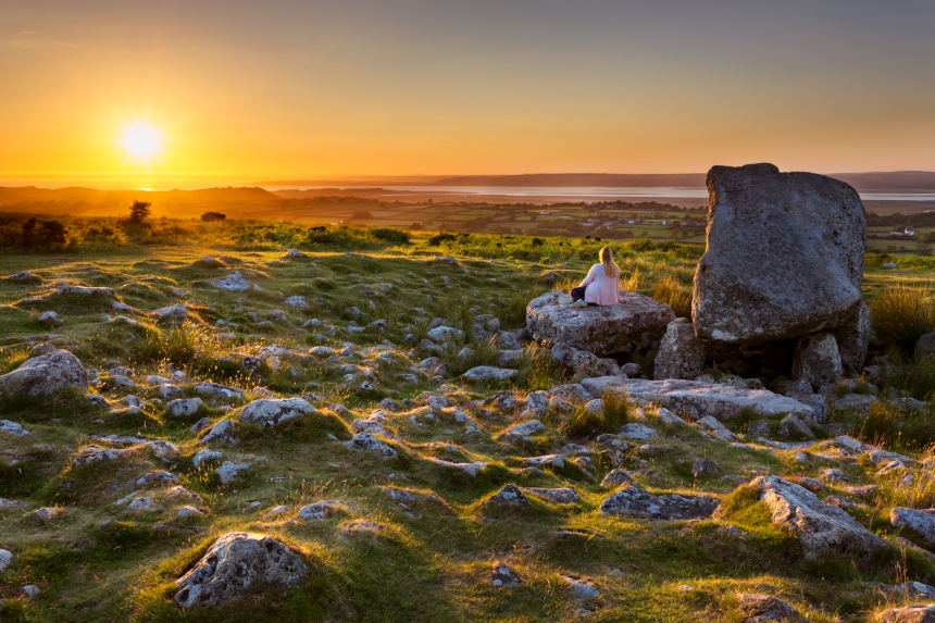 A woman sits on a large limestone rock on Cefn Bryn common on the Gower Peninsula, watching a golden sunset over the Bristol Channel, with rolling green grassland and scattered rocks stretching into the foreground
