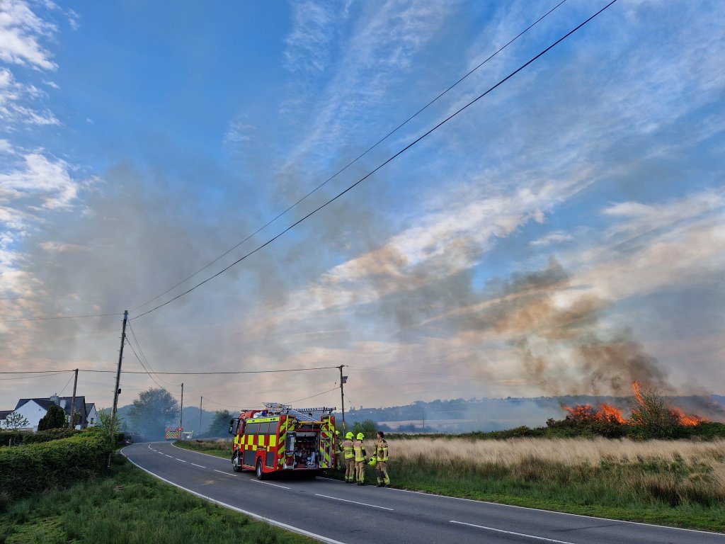A Mid and West Wales Fire and Rescue Service fire engine parked on a country road on Gower with firefighters standing alongside it as flames burn through dry grass and hedgerow beside the road, with a large plume of smoke rising into a blue evening sky