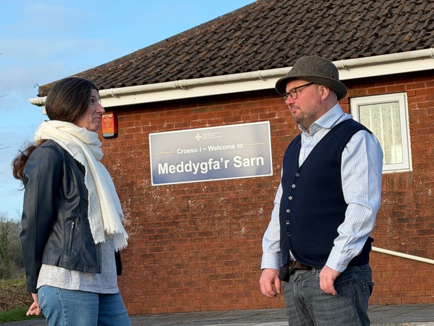 Carl Peters-Bond and Clare Treharne stand outside Meddygfa'r Sarn GP surgery in Pontyates, with the surgery's bilingual welcome sign visible on the wall behind them