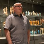 Dr Ken Griffin standing in front of illuminated glass display cases containing rows of ancient Egyptian figurines and sculptures in the Egypt Centre's House of Death gallery.