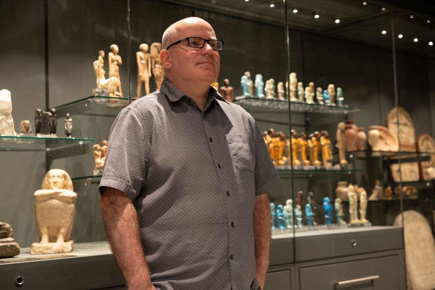 Dr Ken Griffin standing in front of illuminated glass display cases containing rows of ancient Egyptian figurines and sculptures in the Egypt Centre's House of Death gallery.