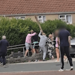 A person raises a large wooden plank above their head during a street brawl in a residential area of Swansea, with several other individuals visible around them and a red-roofed house in the background. Some faces have been blurred.