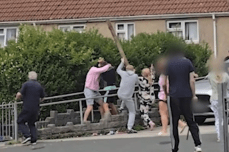 A person raises a large wooden plank above their head during a street brawl in a residential area of Swansea, with several other individuals visible around them and a red-roofed house in the background. Some faces have been blurred.