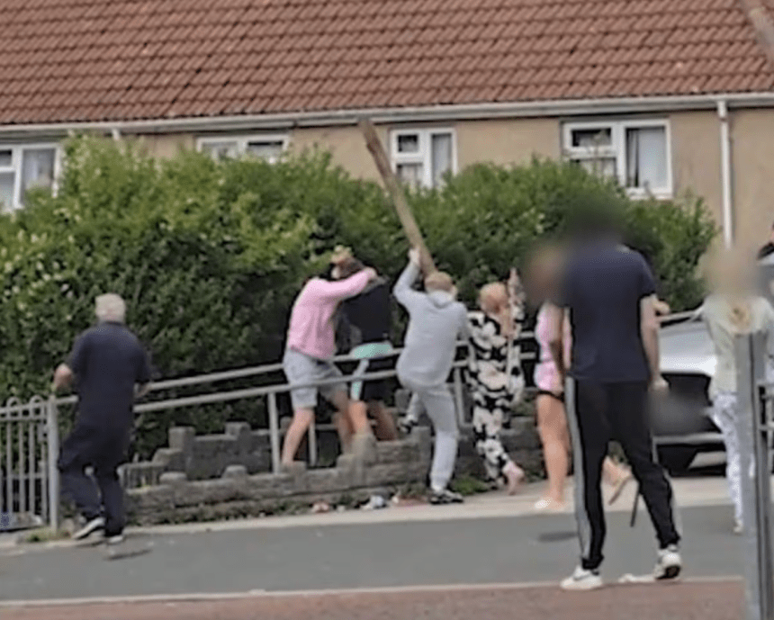 A person raises a large wooden plank above their head during a street brawl in a residential area of Swansea, with several other individuals visible around them and a red-roofed house in the background. Some faces have been blurred.