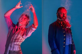 Promotional image for Amadeus showing Callum Scott Howells as Mozart on the left and Michael Sheen as Salieri on the right, both lit dramatically in red and blue