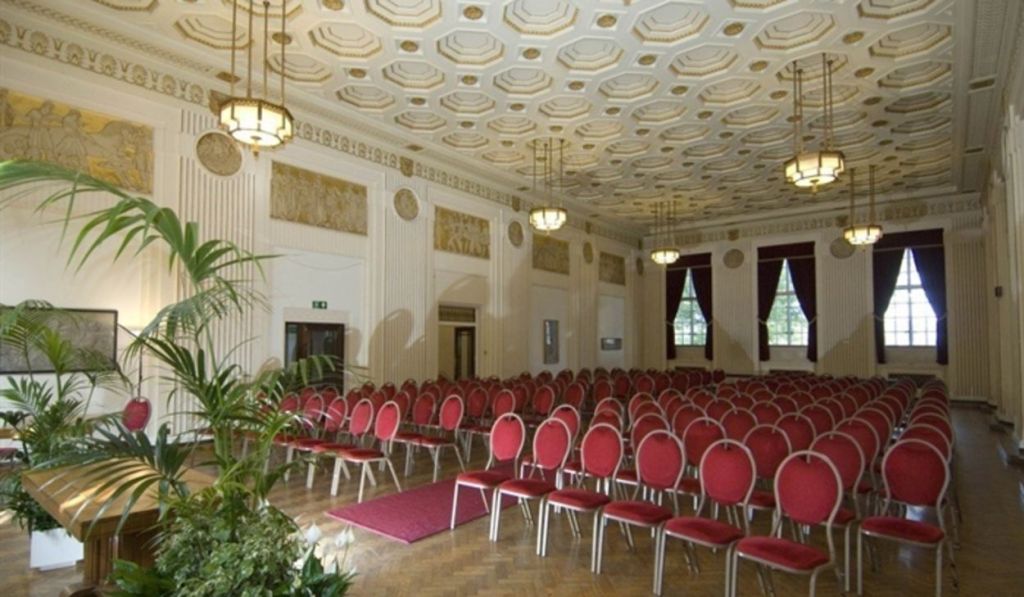 The George Hall at Swansea's Guildhall complex set for a ceremony, showing the ornate coffered ceiling, gold wall friezes and rows of red chairs