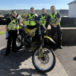 Four South Wales Police officers in high-visibility vests standing with a seized yellow and black off-road motorbike on a residential street in Swansea, with hillside housing visible in the background