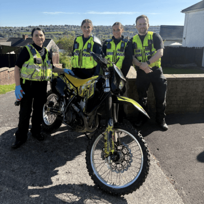 Four South Wales Police officers in high-visibility vests standing with a seized yellow and black off-road motorbike on a residential street in Swansea, with hillside housing visible in the background