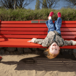 Five-year-old Esme Mariani laughing upside down on a red British Heart Foundation bench on Swansea Beach.