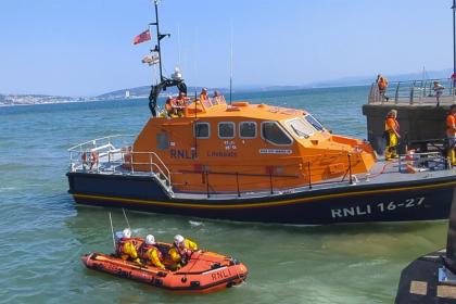The orange Mumbles RNLI all-weather lifeboat Roy Barker IV moored at Mumbles Pier on a sunny day, with the smaller orange inshore lifeboat alongside and crew members on deck, Swansea Bay visible in the background.
