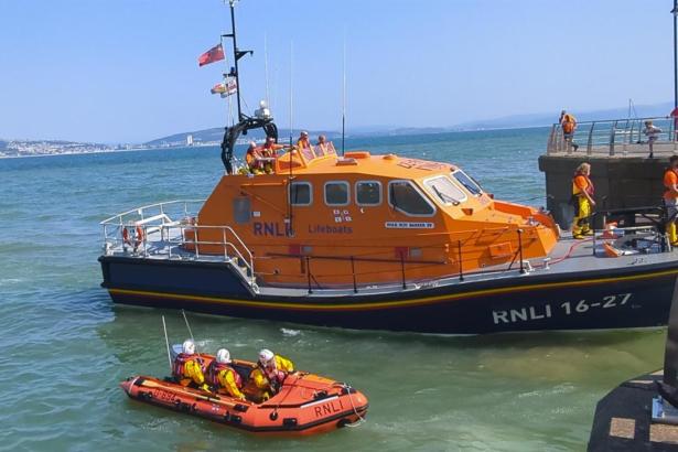 The orange Mumbles RNLI all-weather lifeboat Roy Barker IV moored at Mumbles Pier on a sunny day, with the smaller orange inshore lifeboat alongside and crew members on deck, Swansea Bay visible in the background.