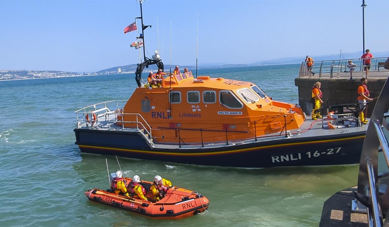 The orange Mumbles RNLI all-weather lifeboat Roy Barker IV moored at Mumbles Pier on a sunny day, with the smaller orange inshore lifeboat alongside and crew members on deck, Swansea Bay visible in the background.