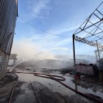 High-pressure water jet arcing over a large pile of smouldering commercial waste at the Port Talbot Dock Road industrial fire, with fire hoses on flooded ground