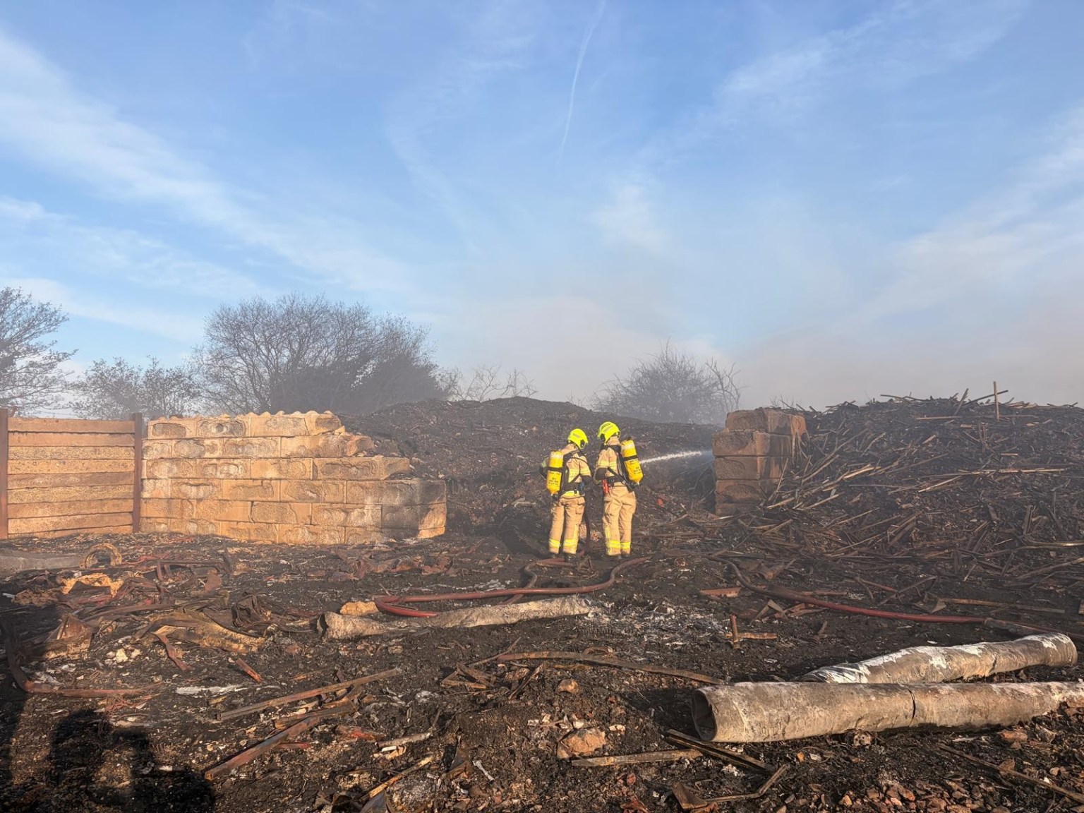 Two firefighters in yellow protective gear and breathing apparatus directing a hose into smouldering debris at the Port Talbot Dock Road fire site