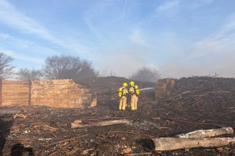 Two firefighters in yellow protective gear and breathing apparatus directing a hose into smouldering debris at the Port Talbot Dock Road fire site