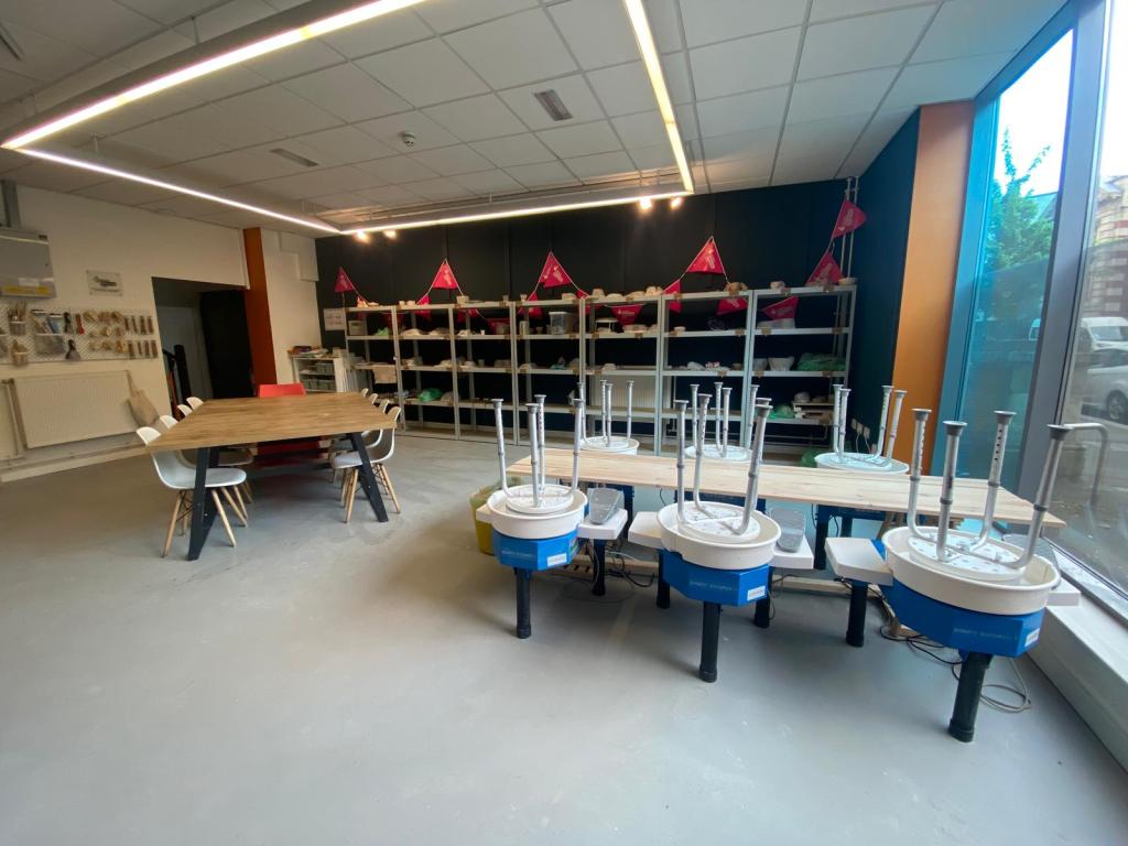 The interior of the Swansea Pottery Collective studio, showing a row of blue electric pottery wheels on a wooden workbench, a large oak dining table with chairs, shelving stocked with equipment and clay, and large windows overlooking the street.