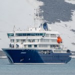 The MV Hondius, a dark blue and white expedition cruise ship, seen from the bow with the name Hondius visible on the hull, sailing in calm grey-blue waters with a snow-covered rocky hillside behind it.