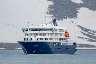 The MV Hondius, a dark blue and white expedition cruise ship, seen from the bow with the name Hondius visible on the hull, sailing in calm grey-blue waters with a snow-covered rocky hillside behind it.