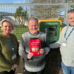 Three people standing outside next to a green wall-mounted defibrillator cabinet, with the man in the middle holding up a red Public Access Bleed Control Kit emergency bag.