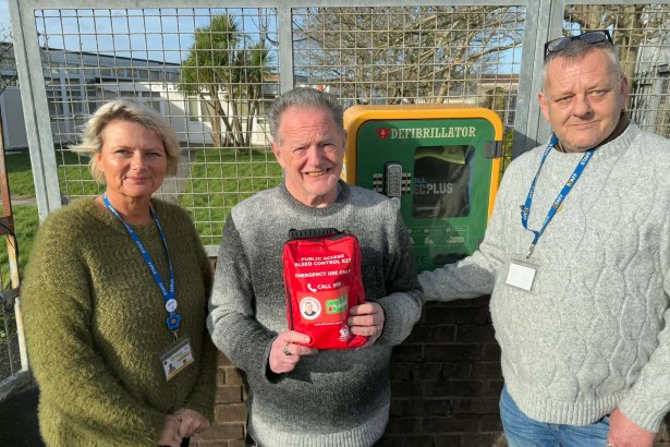 Three people standing outside next to a green wall-mounted defibrillator cabinet, with the man in the middle holding up a red Public Access Bleed Control Kit emergency bag.