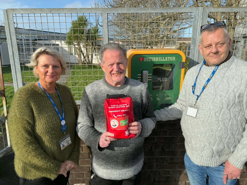 Three people standing outside next to a green wall-mounted defibrillator cabinet, with the man in the middle holding up a red Public Access Bleed Control Kit emergency bag.
