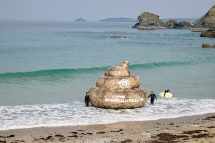 A giant inflatable in the shape of a poo on a beach, branded with the Surfers Against Sewage logo and the hashtag #EndSewagePollution, with surfers standing nearby in the shallows.