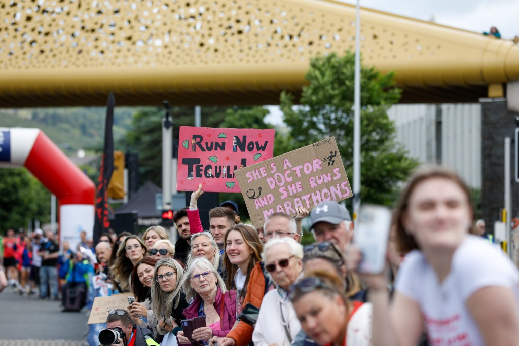 Crowd of spectators standing under the Copr Bay Bridge in Swansea at a previous Mizuno Swansea Half Marathon, holding handmade placards including one reading "Run Now Tequila Later" and another reading "She's a Doctor and She Runs Marathons".