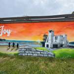 A mural of three children silhouetted running on a beach toward the sea at sunset, with a modernist white building on the right and an orange and blue sky above