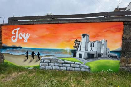 A mural of three children silhouetted running on a beach toward the sea at sunset, with a modernist white building on the right and an orange and blue sky above