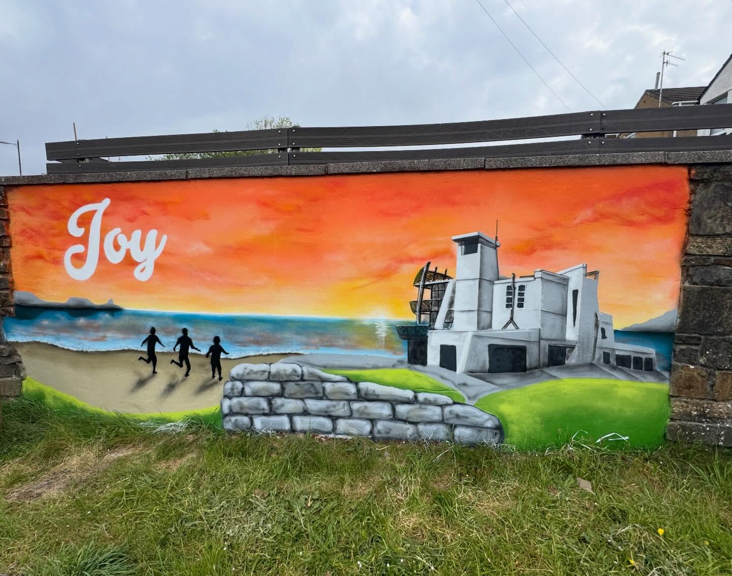 A mural of three children silhouetted running on a beach toward the sea at sunset, with a modernist white building on the right and an orange and blue sky above