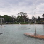 A shallow outdoor lido pool at Blackpill, Swansea, with water jets and sprinklers in operation, playground equipment visible in the background and trees and cloudy sky above