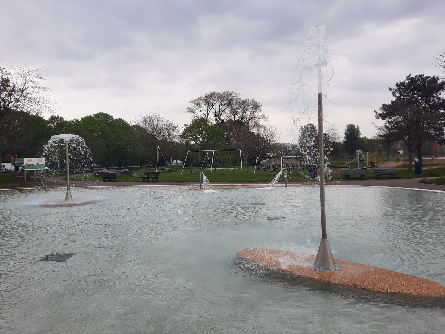 The shallow outdoor paddling pool at Blackpill Lido with its water jets and sprinklers running, playground equipment and trees in the background