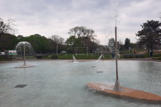 A shallow outdoor lido pool at Blackpill, Swansea, with water jets and sprinklers in operation, playground equipment visible in the background and trees and cloudy sky above