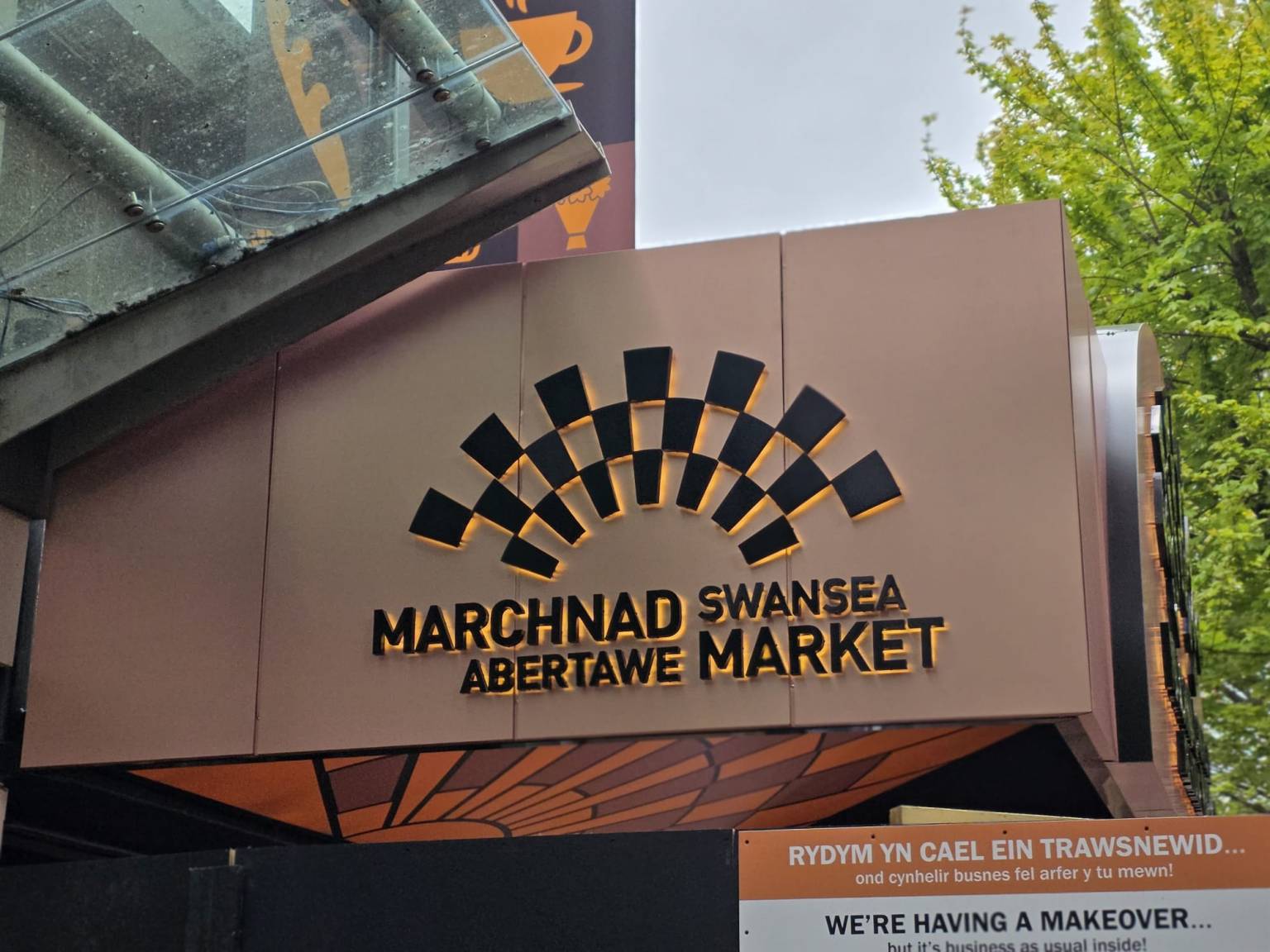 Close-up of the new illuminated Marchnad Abertawe Swansea Market sign at the Union Street entrance, showing the distinctive black and gold fan-shaped logo lit up against a bronze-coloured fascia