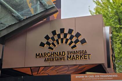 Close-up of the new illuminated Marchnad Abertawe Swansea Market sign at the Union Street entrance, showing the distinctive black and gold fan-shaped logo lit up against a bronze-coloured fascia
